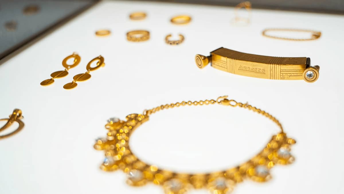 Zwei goldene Schmuckstücke mit runder Scheibe und verziertem Aufsatz liegen nebeneinander auf einer hellen Fläche. Darunter ist eine goldene, konisch geformte Perle in Seitenansicht und in Draufsicht abgebildet.