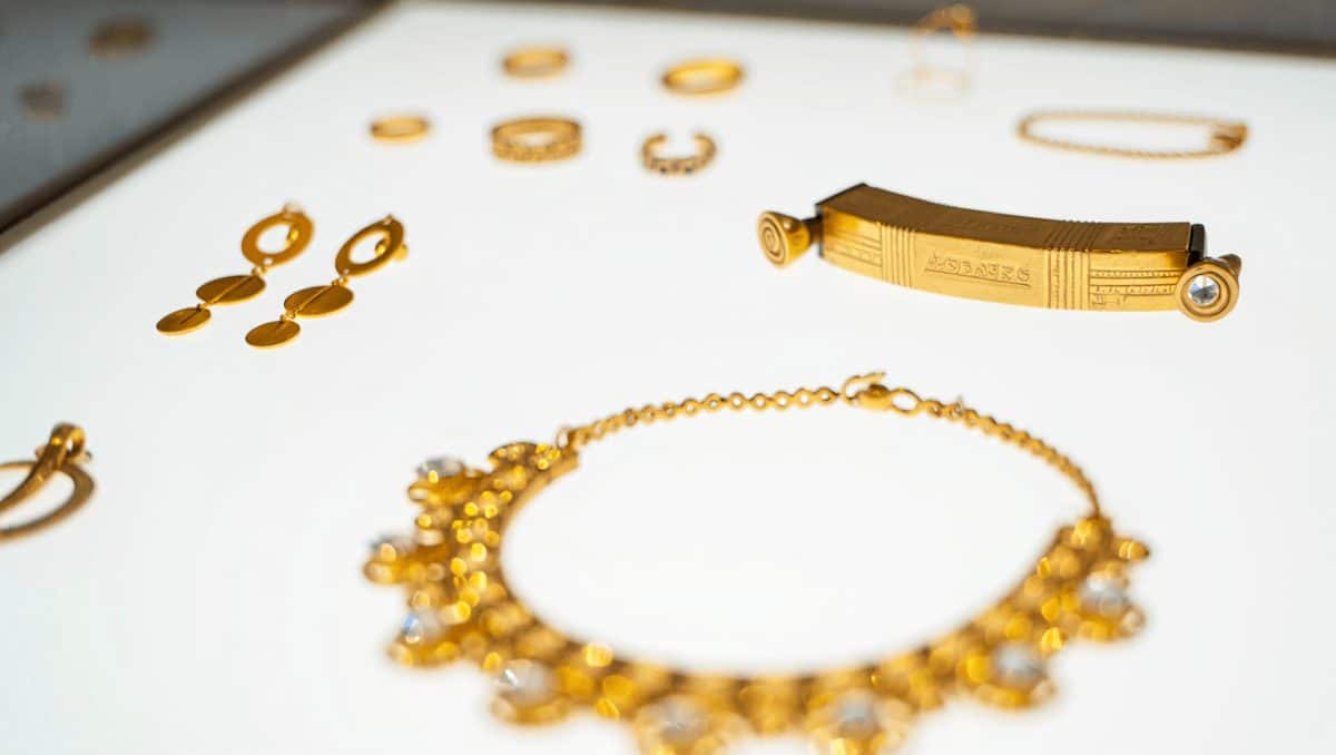 Zwei goldene Schmuckstücke mit runder Scheibe und verziertem Aufsatz liegen nebeneinander auf einer hellen Fläche. Darunter ist eine goldene, konisch geformte Perle in Seitenansicht und in Draufsicht abgebildet.