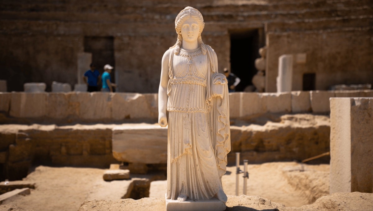 Eine antike Statue liegt auf dem Boden einer Ausgrabungsstätte. Mehrere Personen beugen sich über die Statue und arbeiten gemeinsam daran, sie zu bergen. Die Statue ist aus hellem Stein gefertigt und zeigt eine aufwendig gestaltete Figur mit langen Gewändern. Der Hintergrund besteht aus einer sandfarbenen, steinernen Wand.