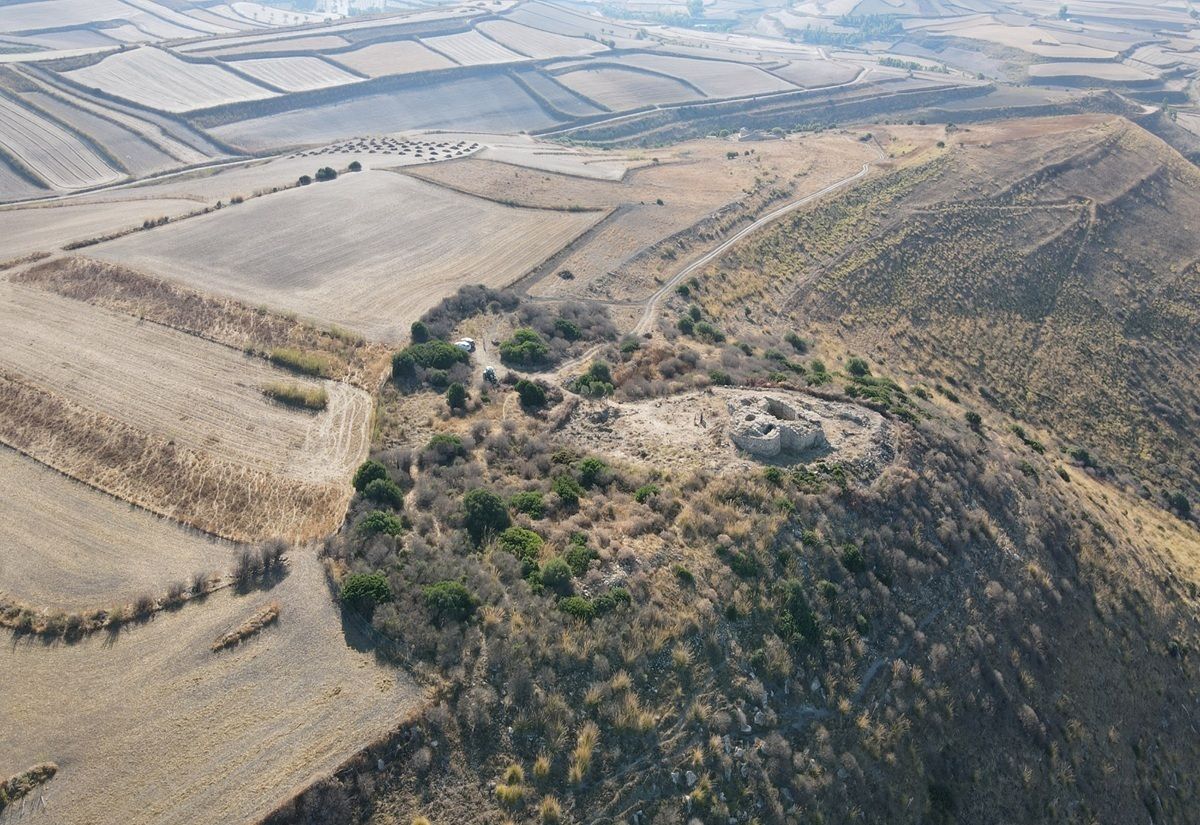 Auf einer Anhöhe liegt eine runde, steinerne Ruine, die von niedriger Vegetation und trockenem Gras umgeben ist. Im Vordergrund erstreckt sich der Hang mit vereinzelten Büschen, während im Hintergrund weite, geometrisch gegliederte Felder zu sehen sind. Die Landschaft wirkt trocken und weitläufig, mit sanften Hügeln und wenigen Bäumen.